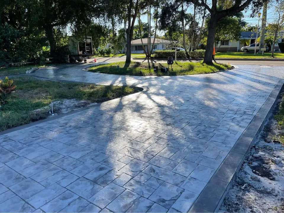 A modern two-story house with a red-tiled roof and white walls is shown. A sleek black car is parked in the paver driveway with high-quality pavers, which includes a small lawn on either side. The house features a carport with columns and tropical landscaping, including palm trees.